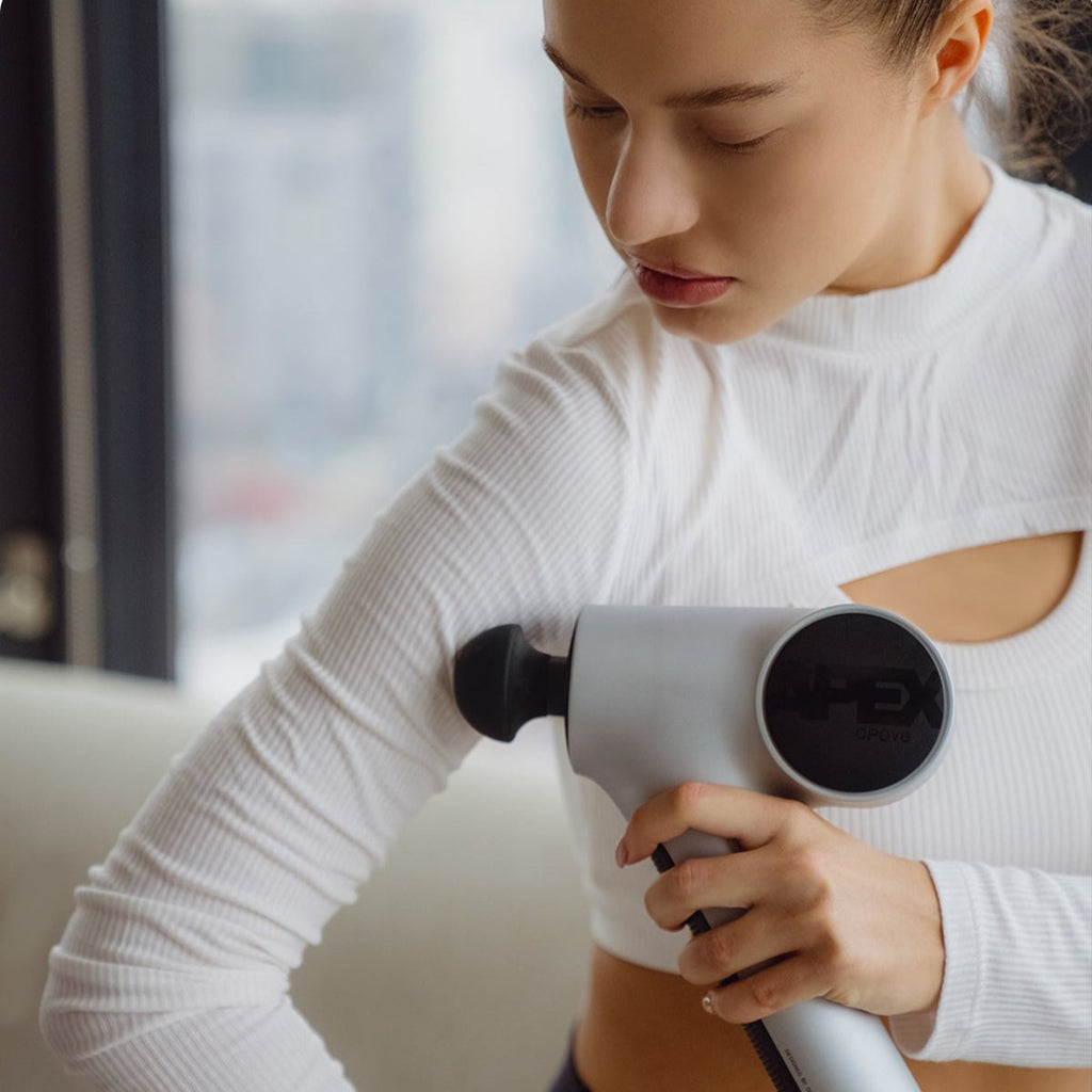 Young woman using the handheld opove Apex Massage Gun—a premium percussive therapy device—to relieve muscle tension in her arm.